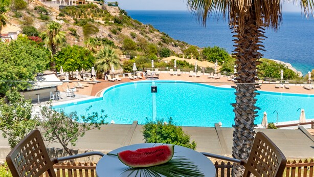 Terraza con vista a una piscina y al mar, con dos sillas y una mesa con sandía.