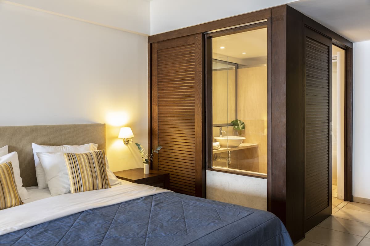 A hotel room with a bed, bedside table, lamp, and a sliding door leading to a bathroom with a sink and mirror.