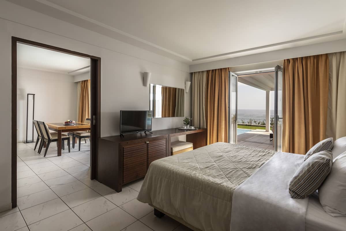 A hotel room with a bed, a desk with a TV, a dining area, and a view of the outdoors.