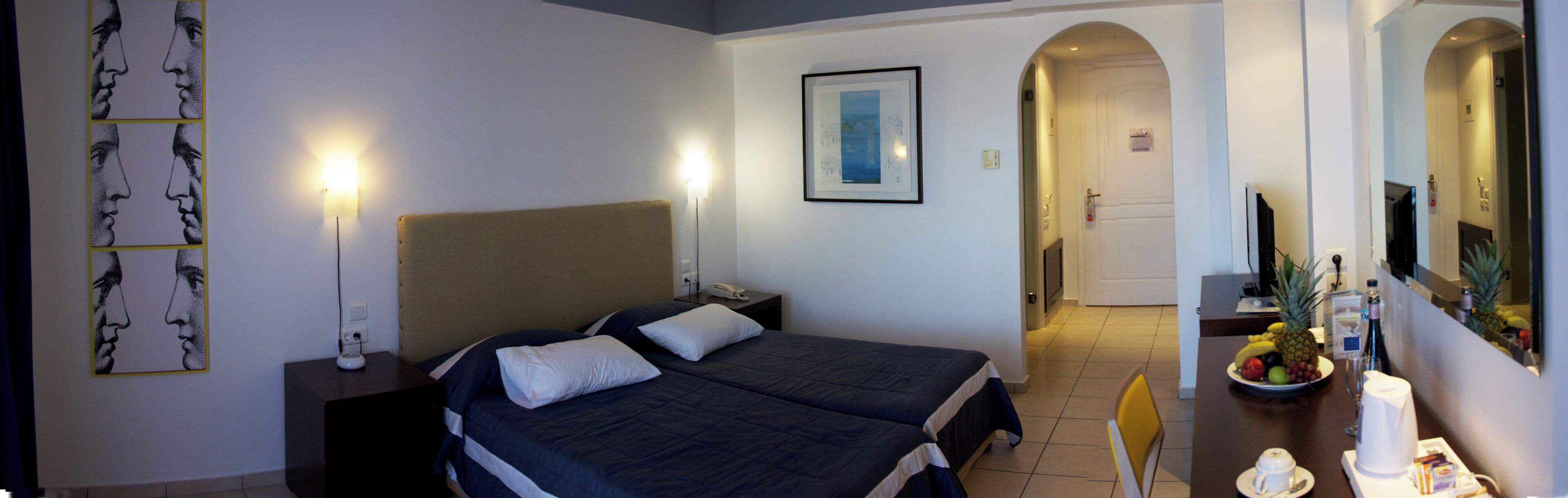 Ein Hotelzimmer mit Bett, Nachttischen, einer Kommode, einem gerahmten Bild an der Wand, einer Tür, die in einen anderen Raum führt, und einem kleinen Tisch mit Obstschale und Kaffeemaschine.