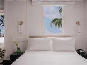 Un dormitorio con una cama, una mesita de noche con teléfono, un florero con flores y una ventana con vista a palmeras.
