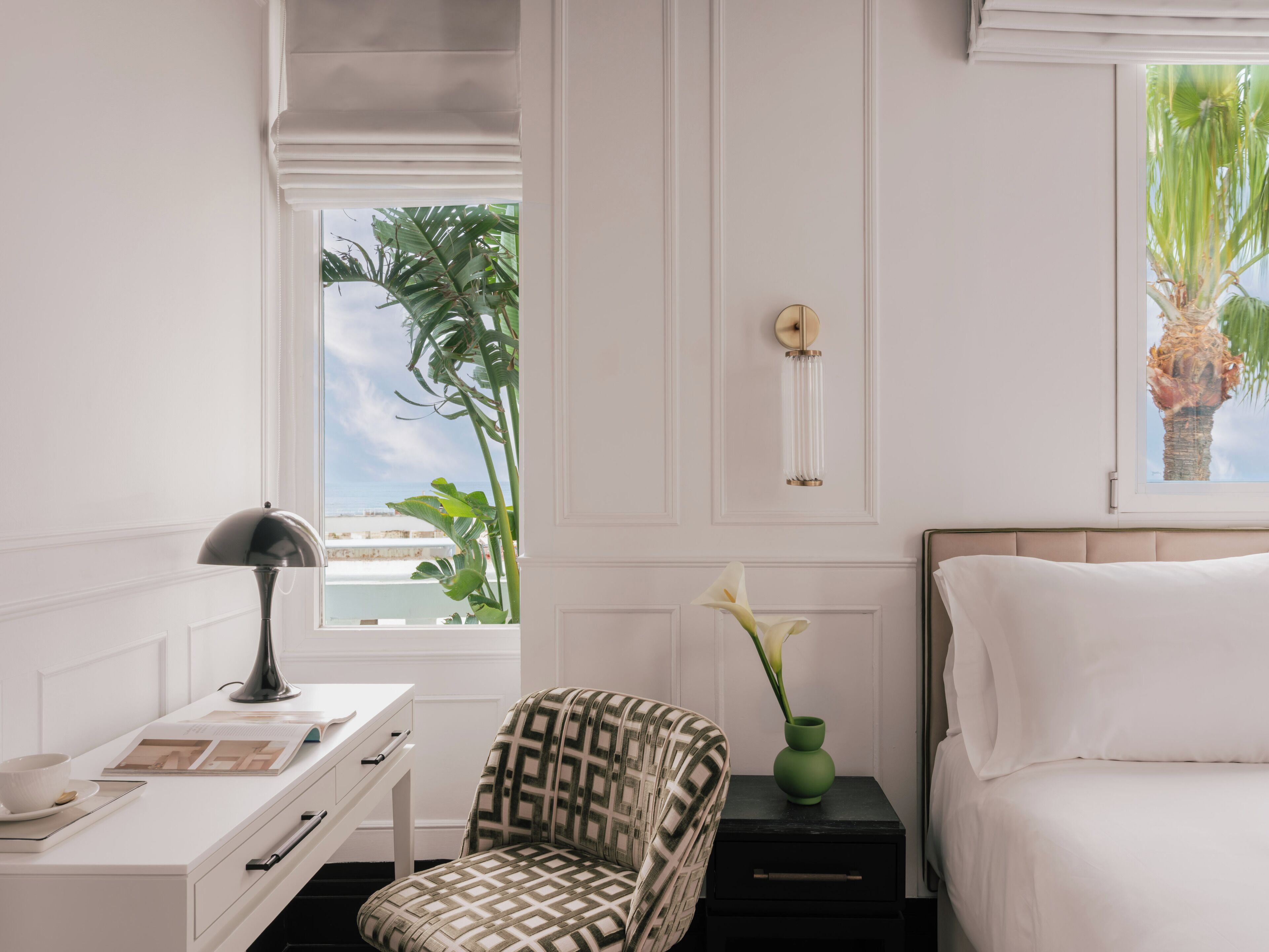 Un dormitorio con cama, escritorio con lámpara, silla, mesita de noche con un florero y ventana con vista a palmeras.