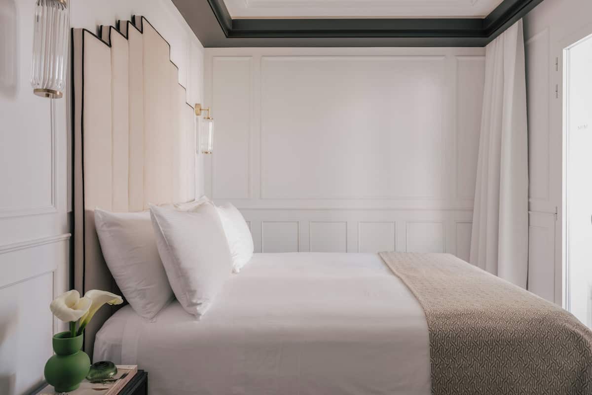 A neatly made bed with white linens, a green vase with flowers, and a book on the nightstand.
