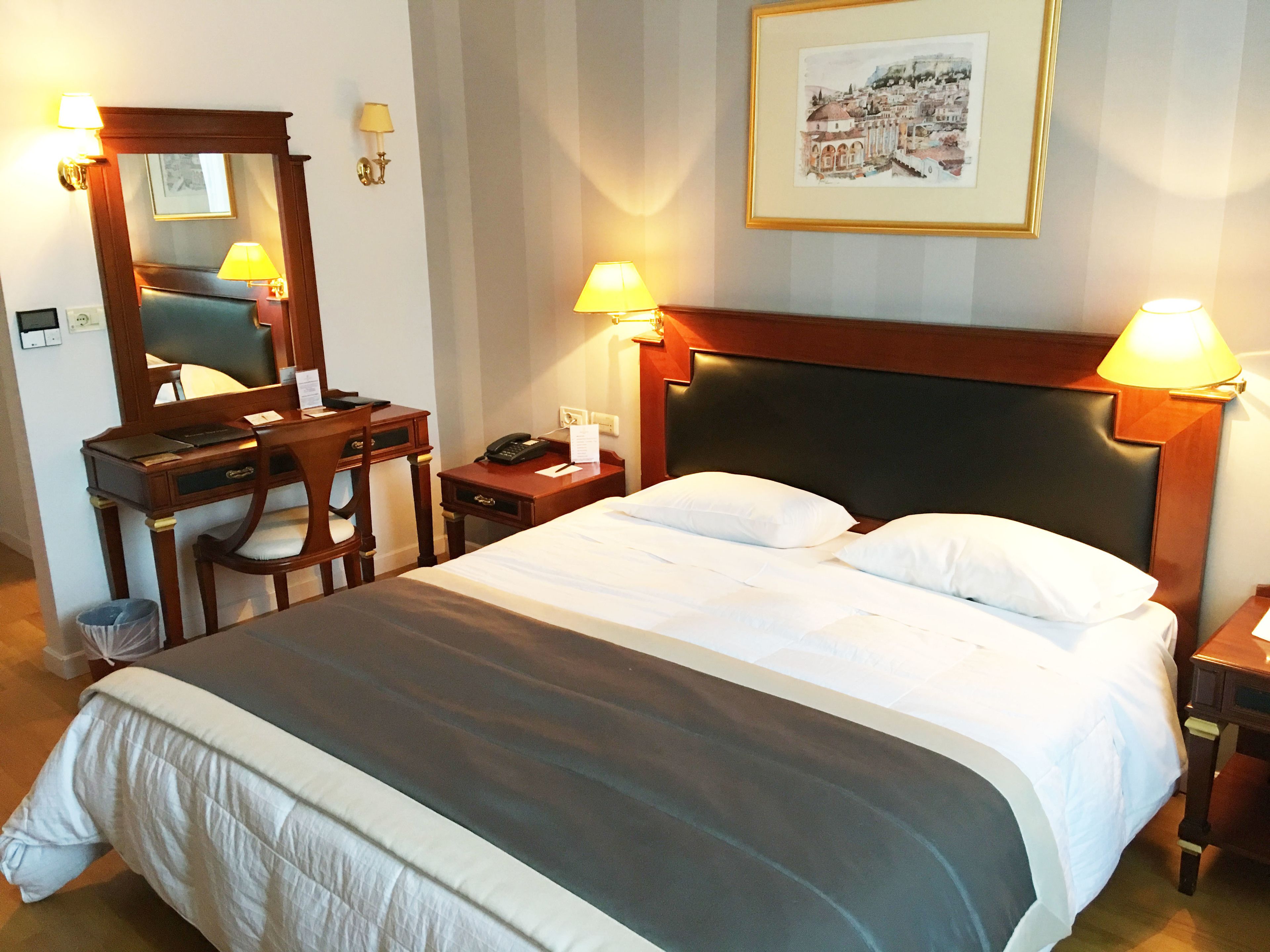 A hotel room with a large bed, a desk with a chair, a desk lamp, a telephone, and a framed picture on the wall.