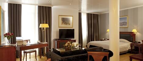 A hotel room with a bed, a desk with a computer, a TV, a dining table with fruit, and a sofa.