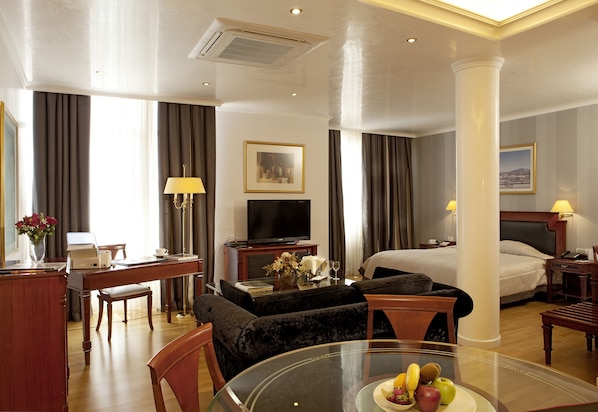 A hotel room with a bed, a desk with a computer, a TV, a dining table with fruit, and a sofa.