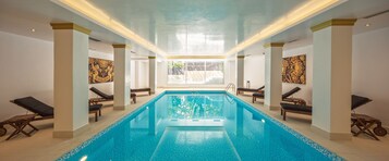 Una piscina interior con agua azul cristalina, rodeada de sillones de descanso y columnas.