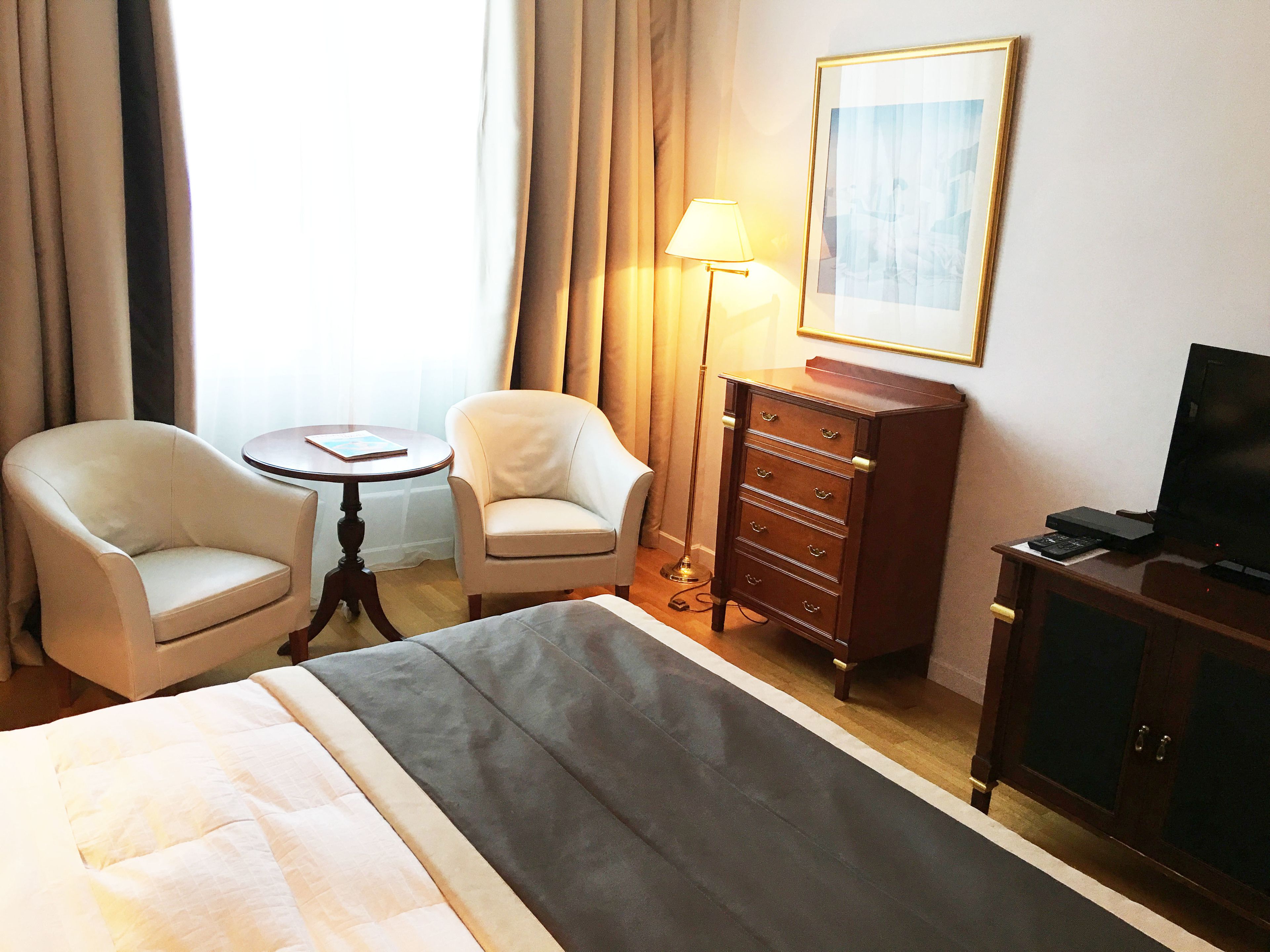 A hotel room with a bed, two armchairs, a small table, a lamp, a dresser, and a television.