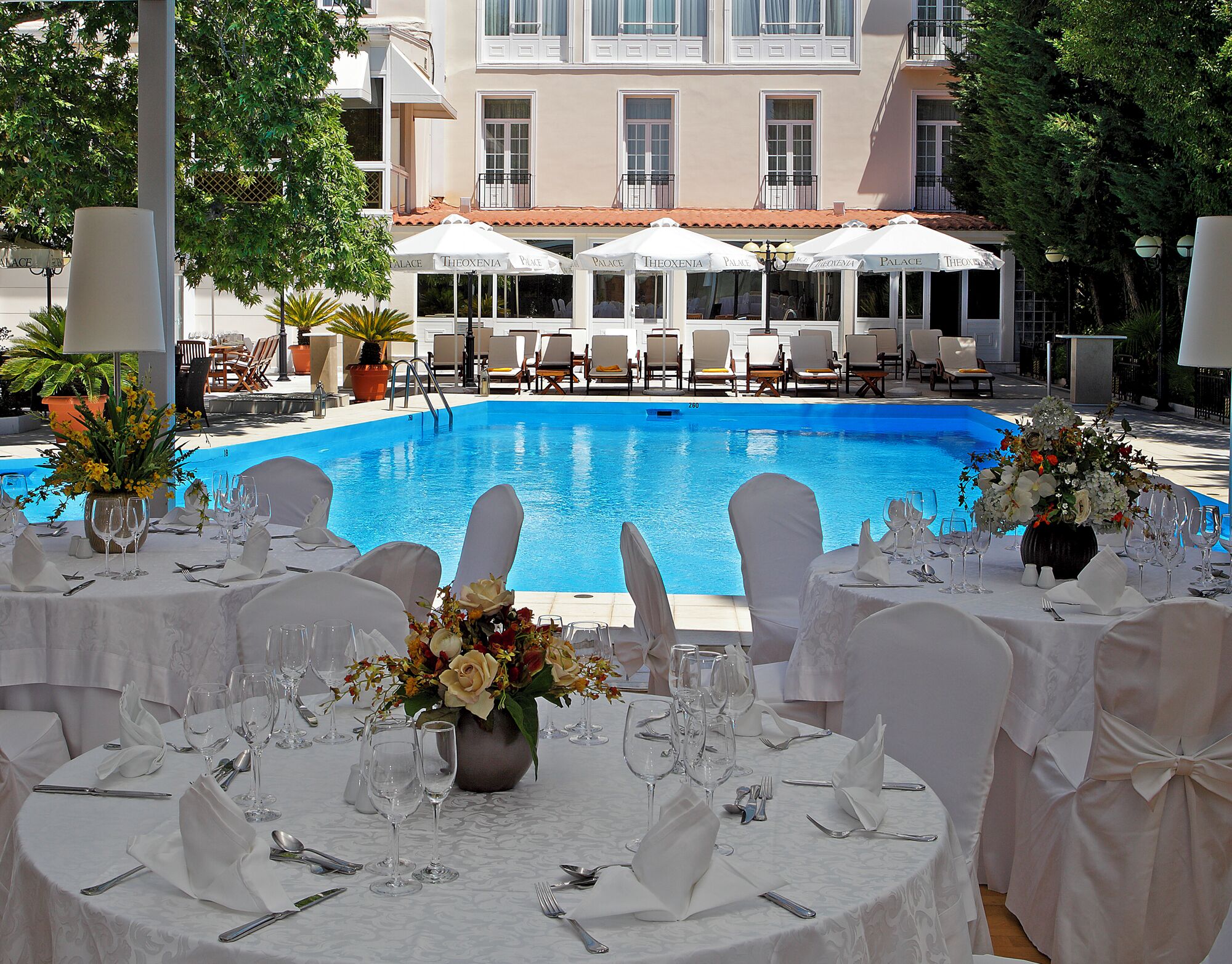 Ein Essbereich am Pool mit Tischen, die für eine festliche Veranstaltung gedeckt sind, mit Blick auf einen Swimmingpool und ein Gebäude mit mehreren Balkonen.
