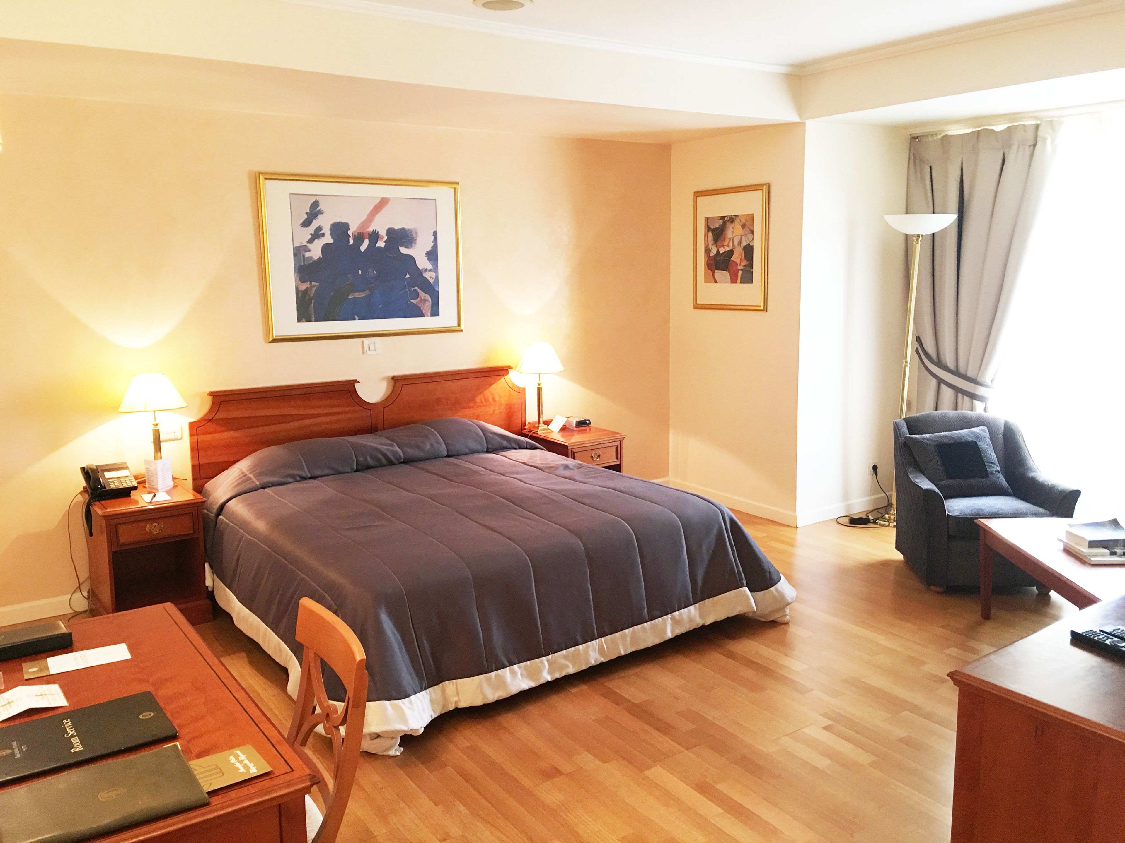 Habitación de hotel con una cama grande, mesitas de noche de madera, un escritorio, una silla y dos cuadros en la pared.