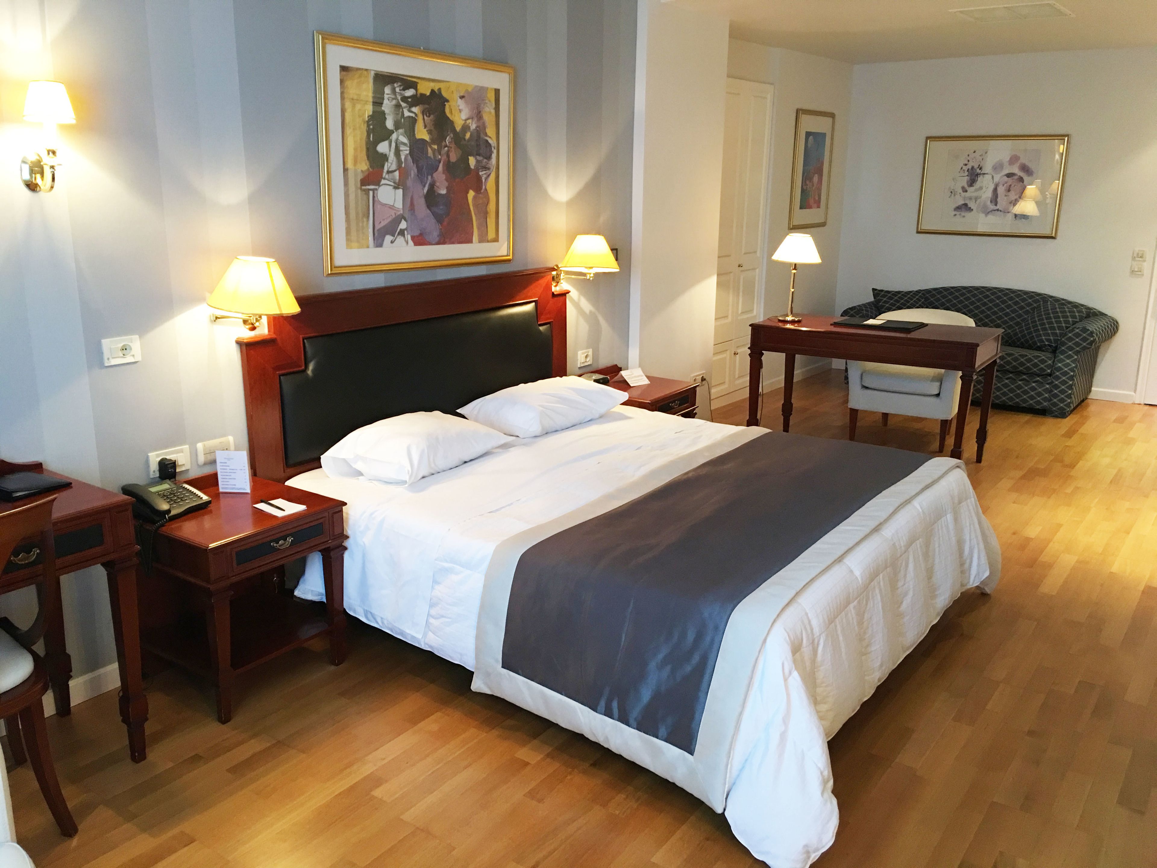 Habitación de hotel con una cama grande, dos mesitas de noche, un escritorio y una silla. Hay dos lámparas en las mesitas de noche y un cuadro en la pared.