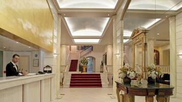 A hotel lobby with a reception desk, a grand staircase, and a decorative mirror.