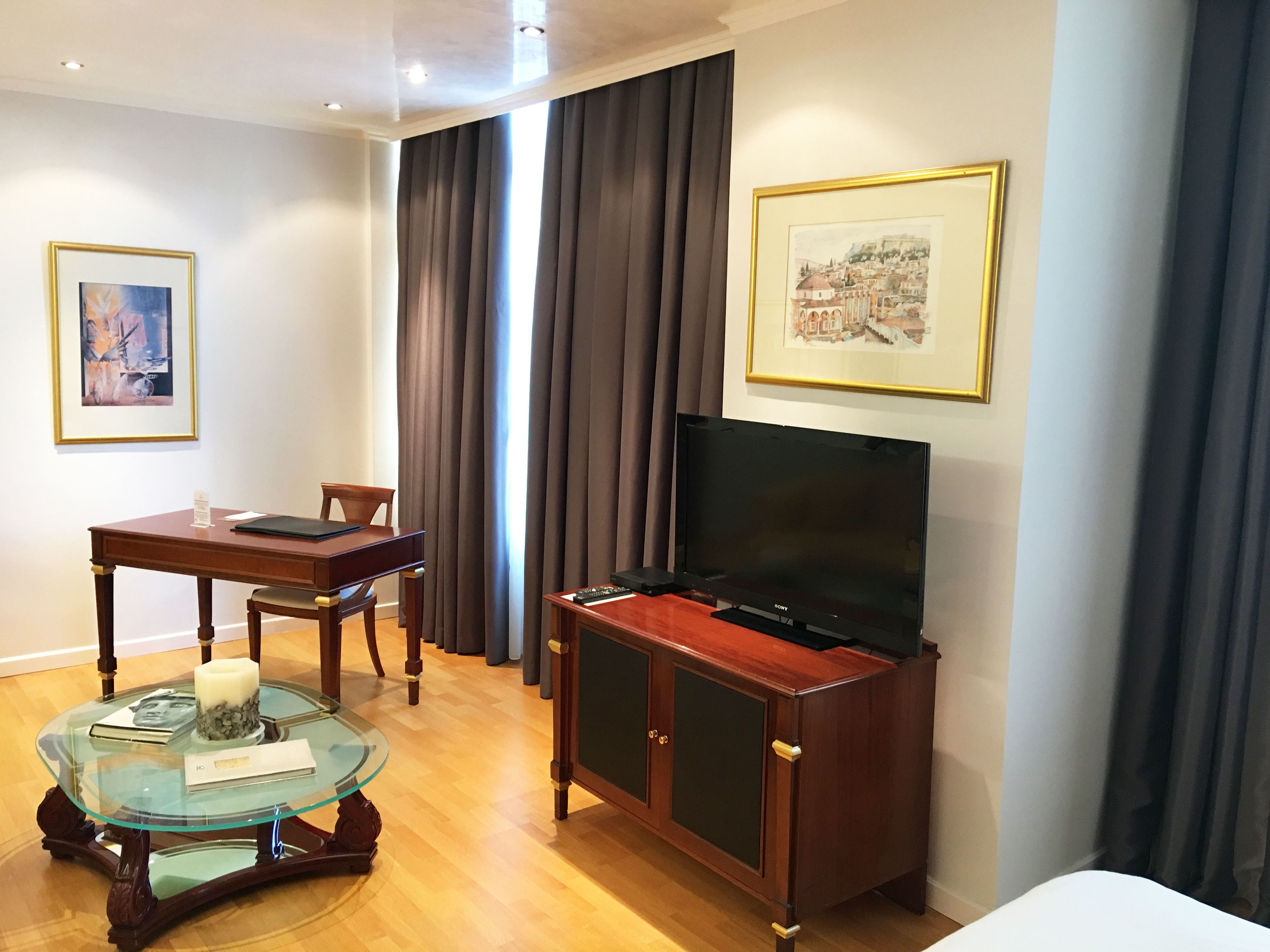 Quarto de hotel com escrivaninha de madeira, mesa de centro de vidro, televisão de tela plana em um armário de madeira e duas fotos emolduradas nas paredes.