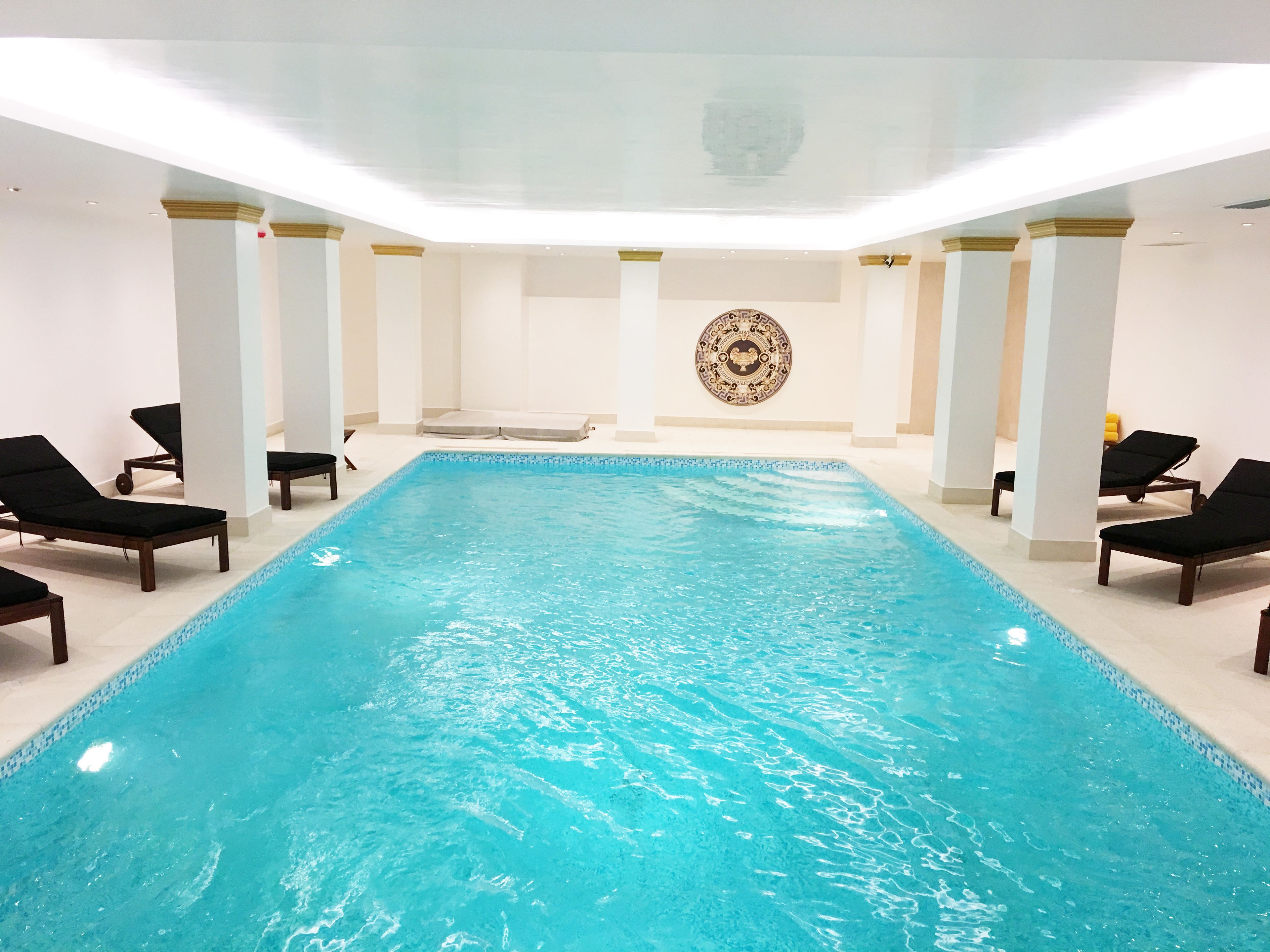 Una piscina interior con agua azul cristalina, rodeada de sillones de descanso y una pared decorativa al fondo.