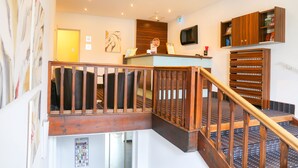 A hotel reception area with a wooden staircase, a reception desk, a mounted television, and a wooden cabinet with shelves. - Hotel Königer (Pliening)