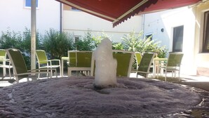 An outdoor seating area with a stone fire pit under a red awning, surrounded by green chairs and tables. - Hotel Königer (Pliening)