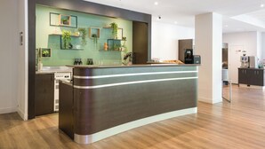 A modern reception desk with a wooden finish, a green wall with plants, and a kitchen area in the background.