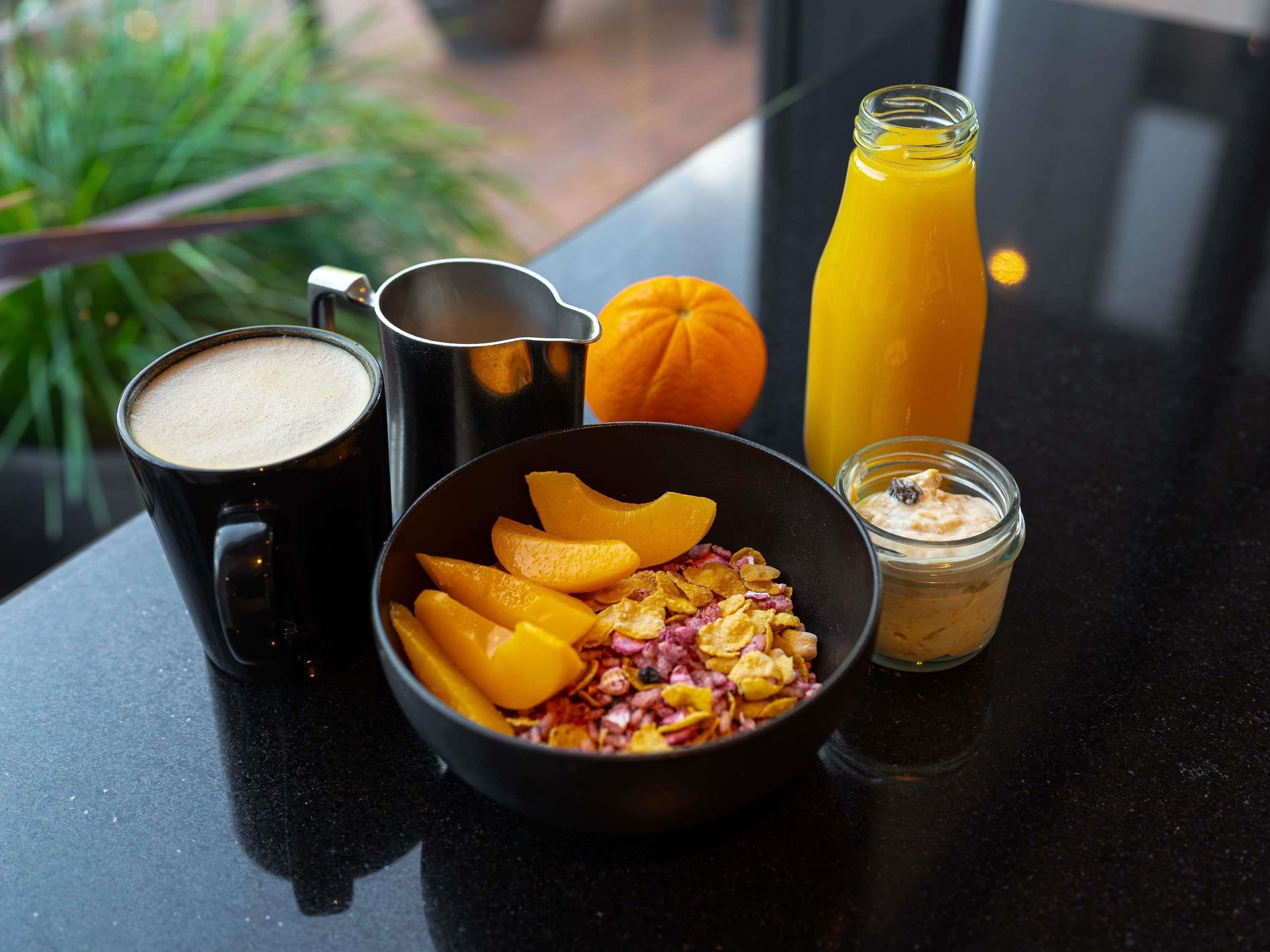 Une table mise pour le petit-déjeuner avec une tasse de café, un bol de fruits et de céréales, un verre de jus d’orange et un petit pot de yaourt.