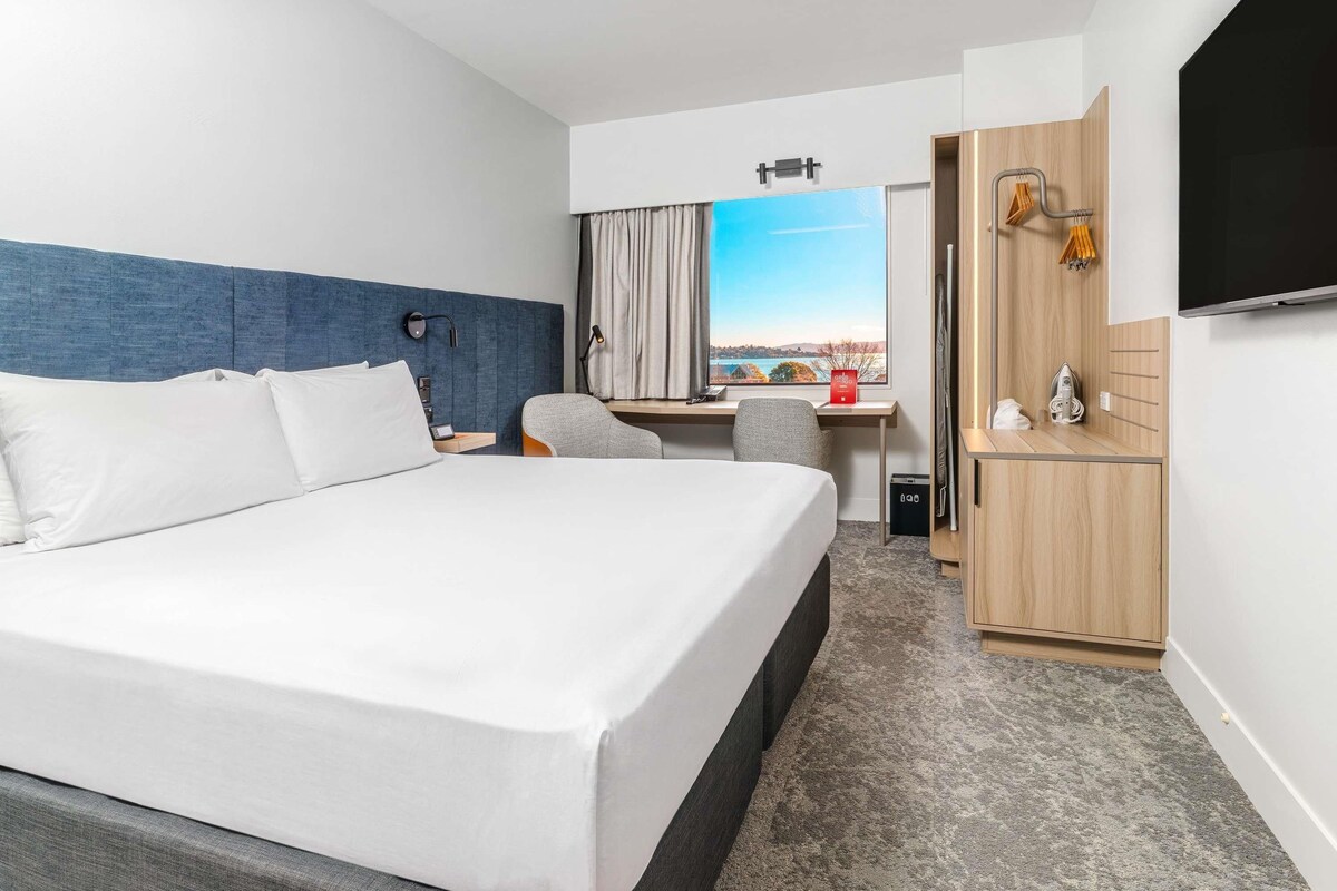 Habitación de hotel con cama, escritorio, silla, televisor y vista al exterior.