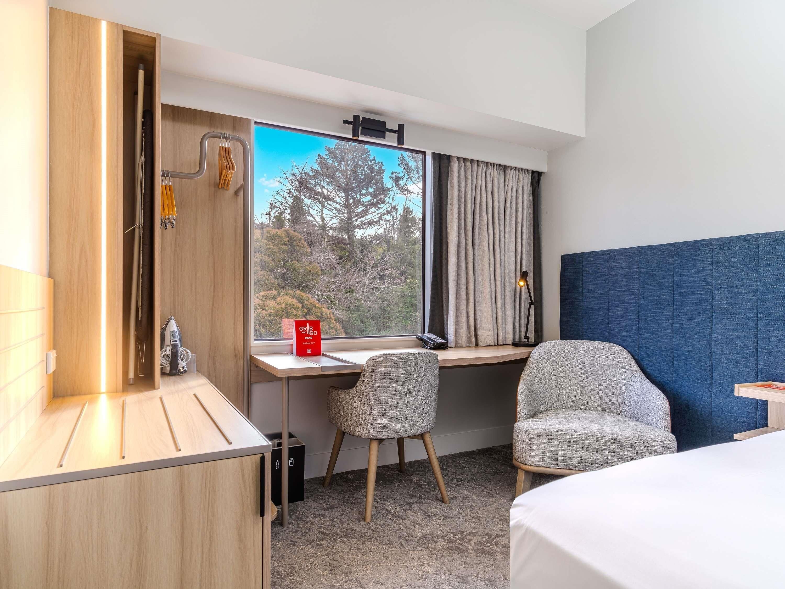 A hotel room with a desk, two chairs, a bed, a window with curtains, and a view of trees.