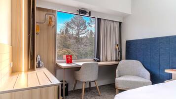 Une chambre d’hôtel équipée d’un bureau, de deux chaises, d’un lit, d’une fenêtre avec des rideaux et offrant une vue sur les arbres.