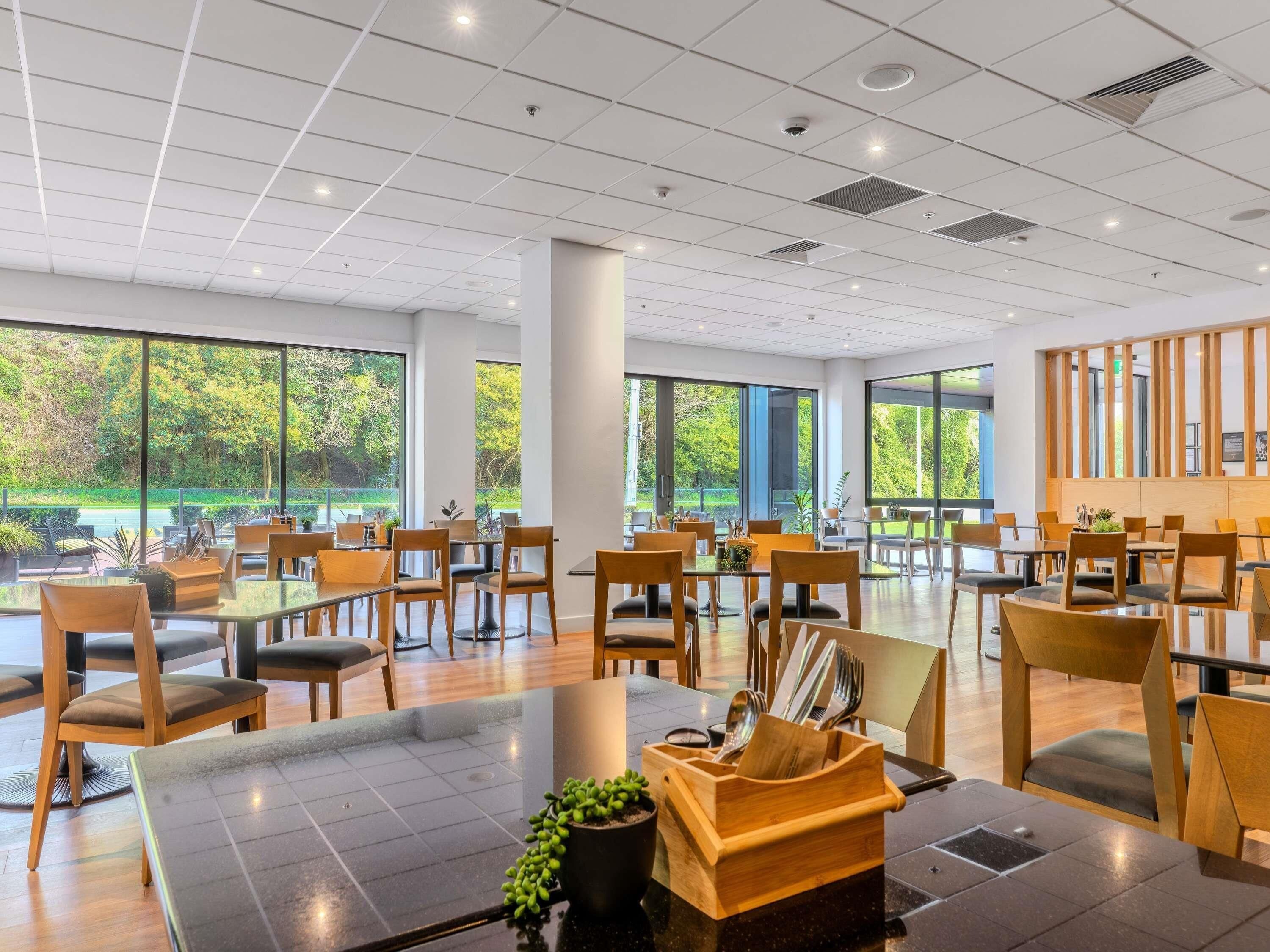 A spacious dining area with multiple tables and chairs, large windows offering views of greenery, and a modern ceiling design.