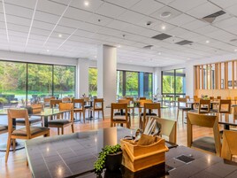 A spacious dining area with multiple tables and chairs, large windows offering views of greenery, and a modern ceiling design.