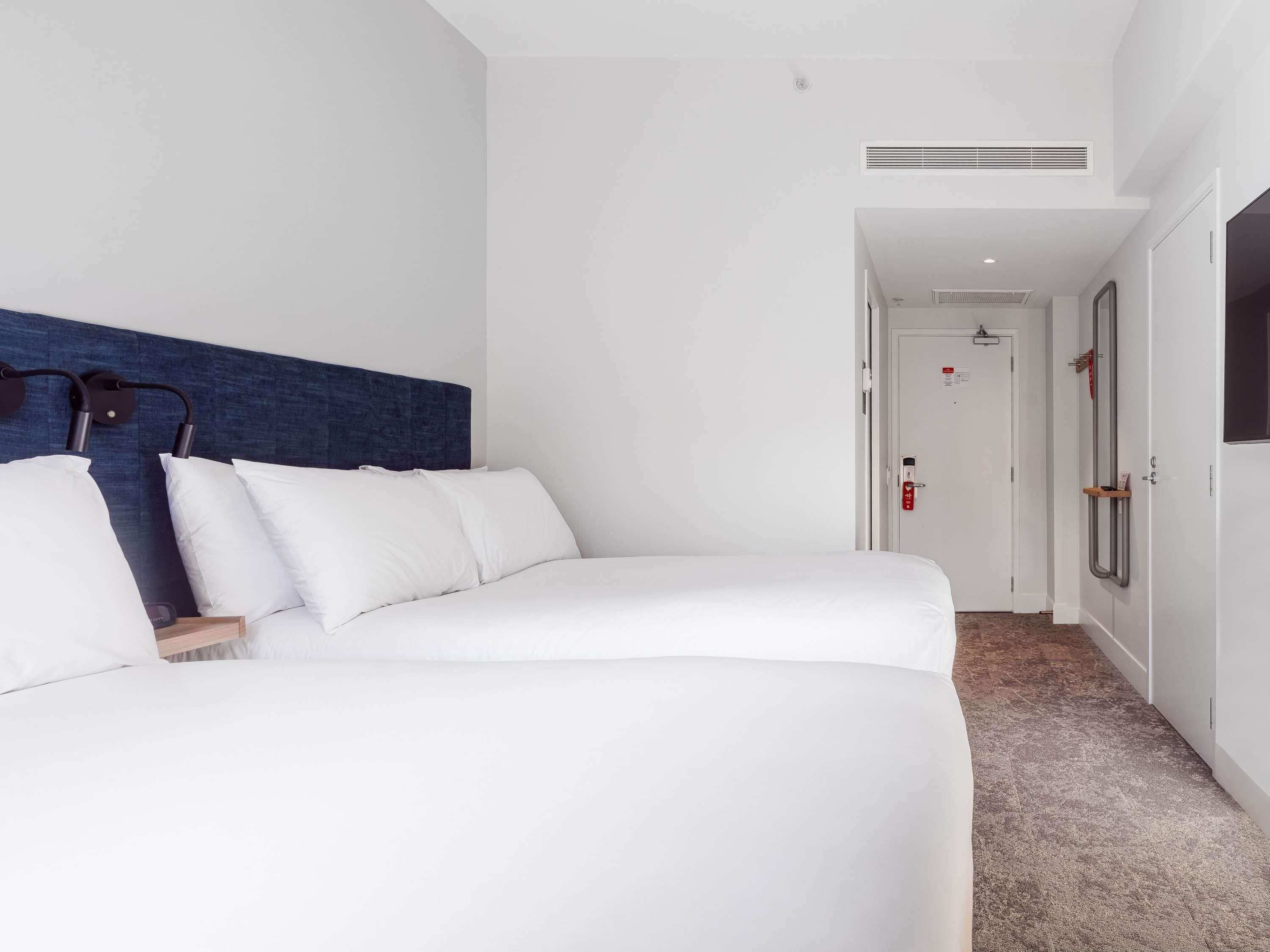 A hotel room with a large bed, a headboard, a bedside table, a nightstand, a wall-mounted lamp, a door with a handle, a window, and a carpeted floor.