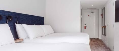 A hotel room with a large bed, a headboard, a bedside table, a nightstand, a wall-mounted lamp, a door with a handle, a window, and a carpeted floor.