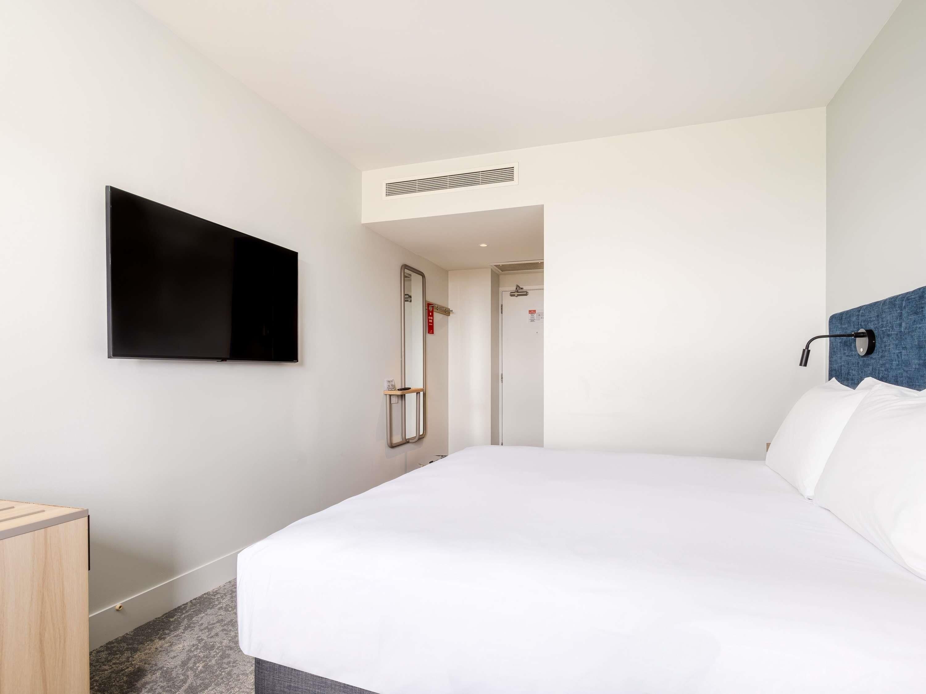 A hotel room with a bed, a television mounted on the wall, a door, and a small bedside table.