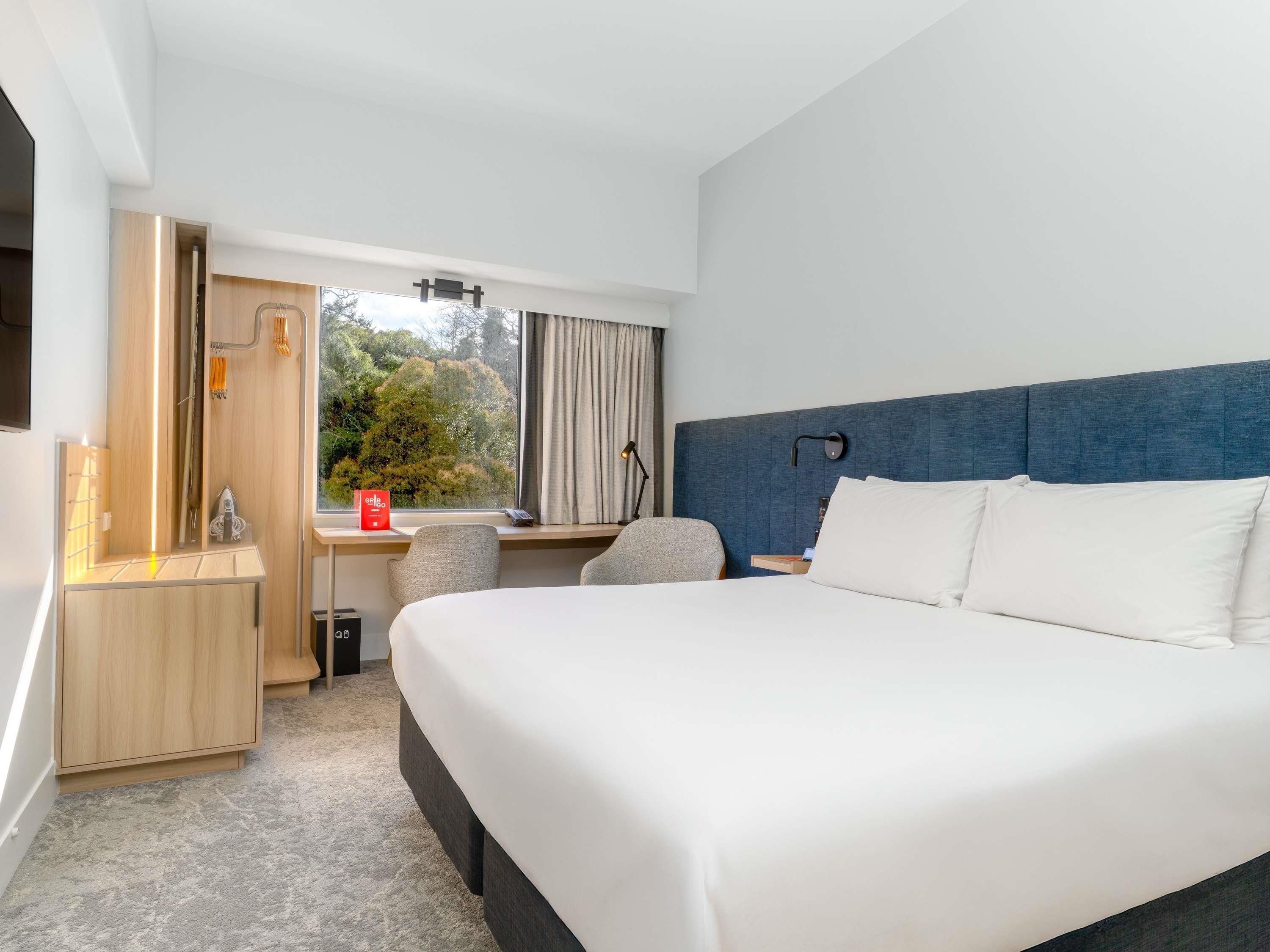 A hotel room with a large bed, a desk, a chair, a window with a view of trees, and a wall-mounted lamp.