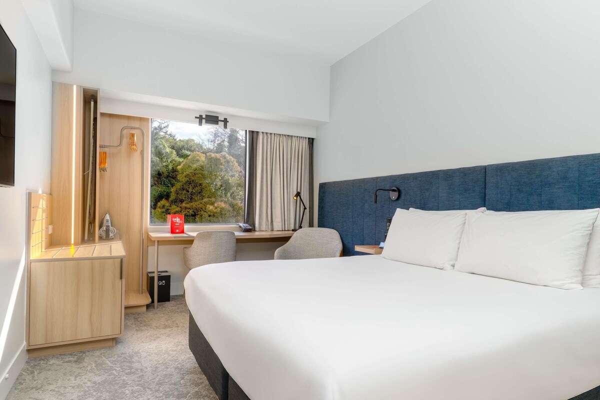 Habitación de hotel con una cama grande, un escritorio, una silla, una ventana con vista a árboles y una lámpara fijada a la pared.