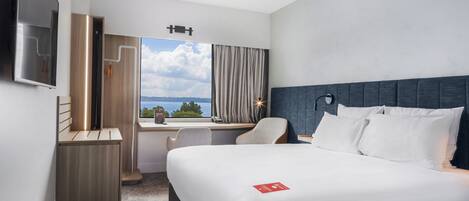 Una habitación de hotel con cama, escritorio, silla, televisor y ventana con vistas al cielo y árboles.
