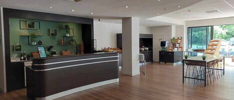 A modern hotel lobby with a reception desk, a dining area, and a kitchen in the background.