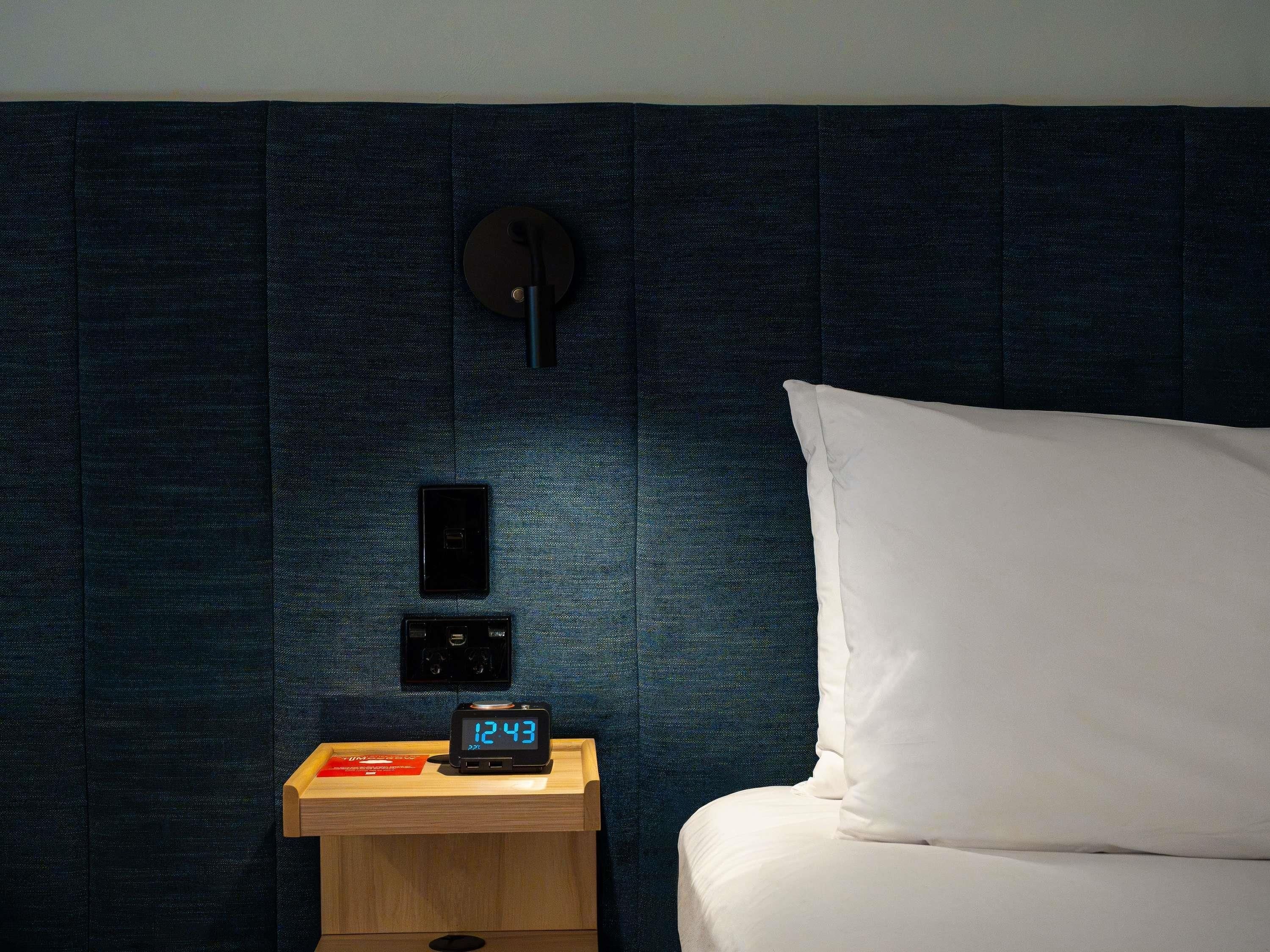 A neatly made bed with a white pillow and a wooden bedside table with a digital clock displaying 12:43.