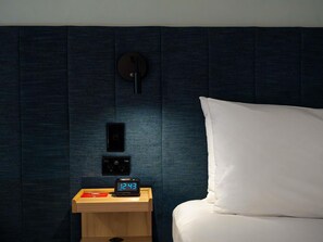A neatly made bed with a white pillow and a wooden bedside table with a digital clock displaying 12:43.