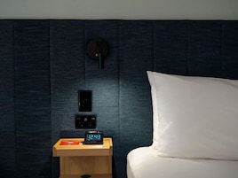 A neatly made bed with a white pillow and a wooden bedside table with a digital clock displaying 12:43.