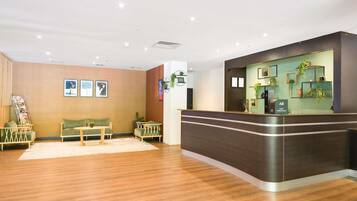 A hotel reception area with a wooden floor, a reception desk, a seating area with a sofa and chairs, and wall-mounted artwork.