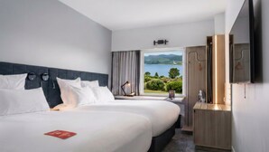 A hotel room with two beds, a large window with a view of the countryside, a flat-screen TV, and a wooden cabinet.
