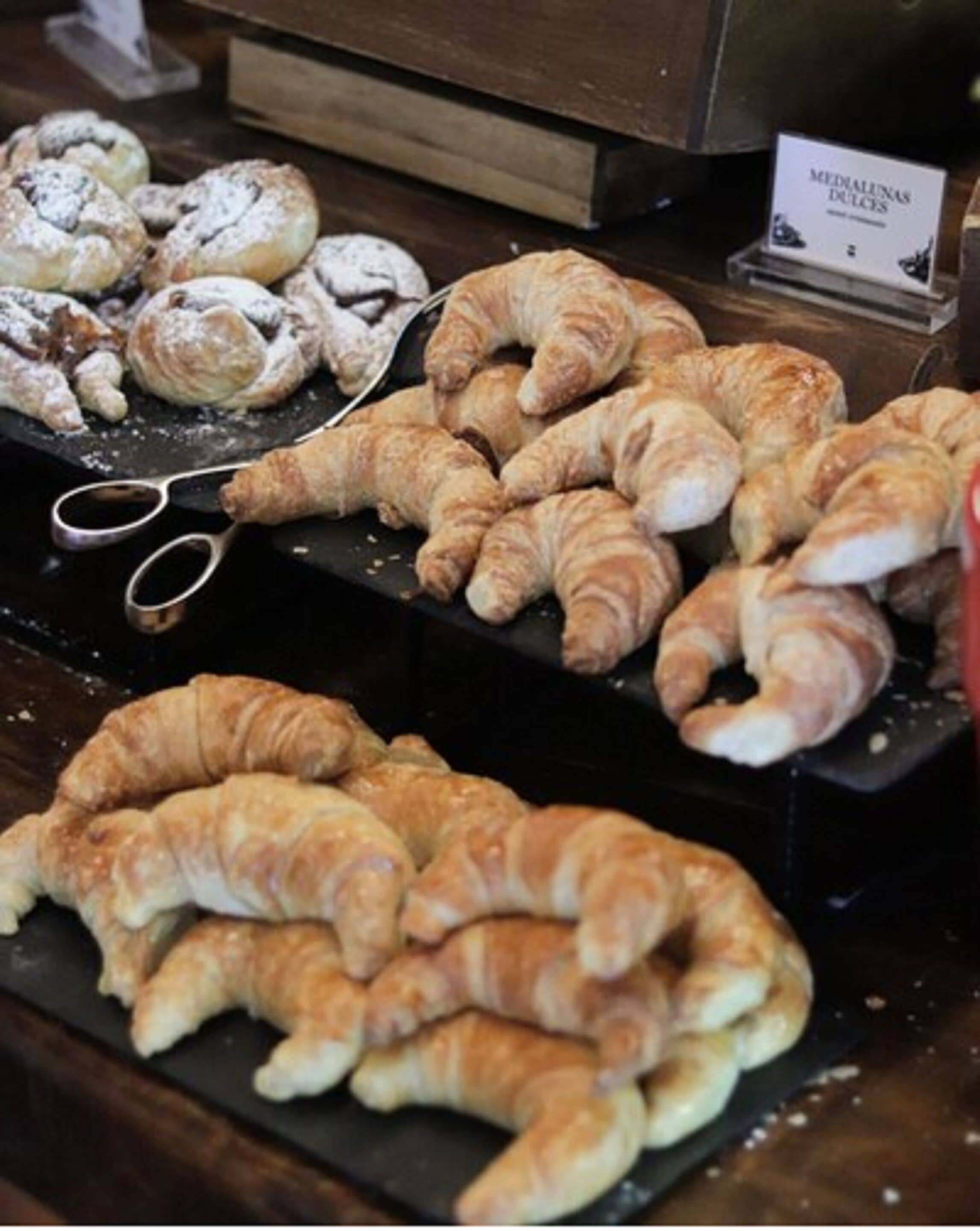 Exposição de diversos doces e pastelaria, incluindo croissants e outros produtos de panificação, dispostos sobre um balcão de madeira.
