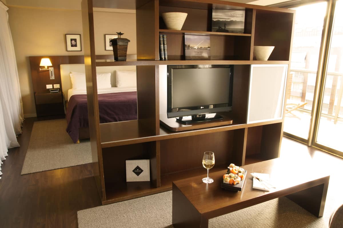 Habitación de hotel con cama, televisión, una mesita de centro con una copa de vino y una estantería.