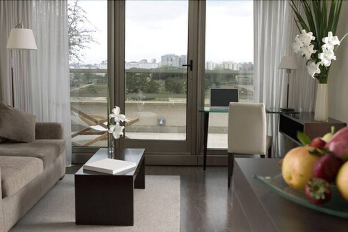 Una sala moderna con un sofá, una mesa de centro y una mesa de vidrio con una computadora portátil. Se aprecia vista de un paisaje urbano a través de amplias ventanas.