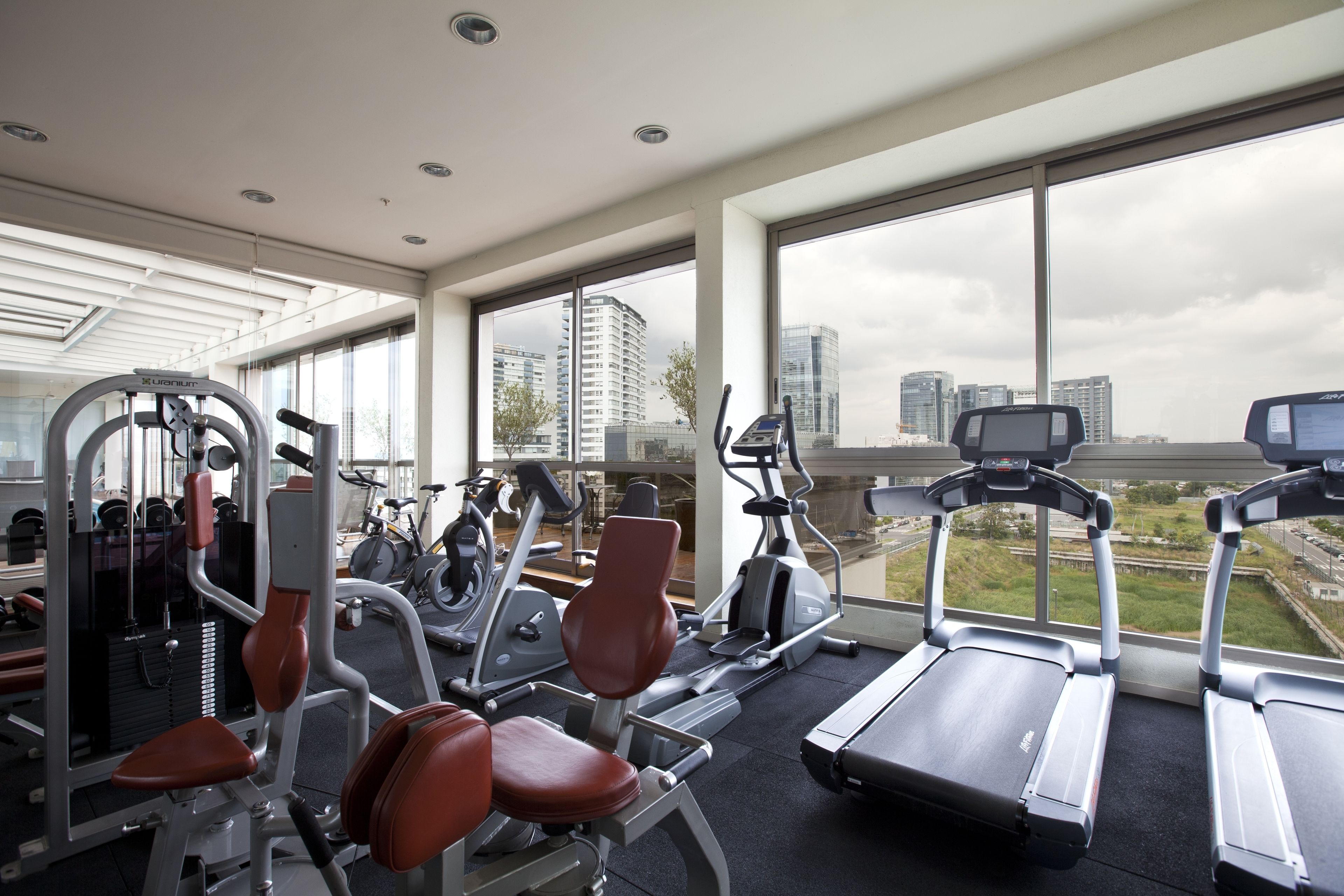 A gym with various exercise equipment, large windows, and a view of a cityscape.