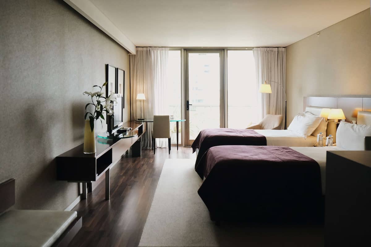 Habitación de hotel con dos camas, un escritorio, una silla, una mesita y un ventanal grande con cortinas.