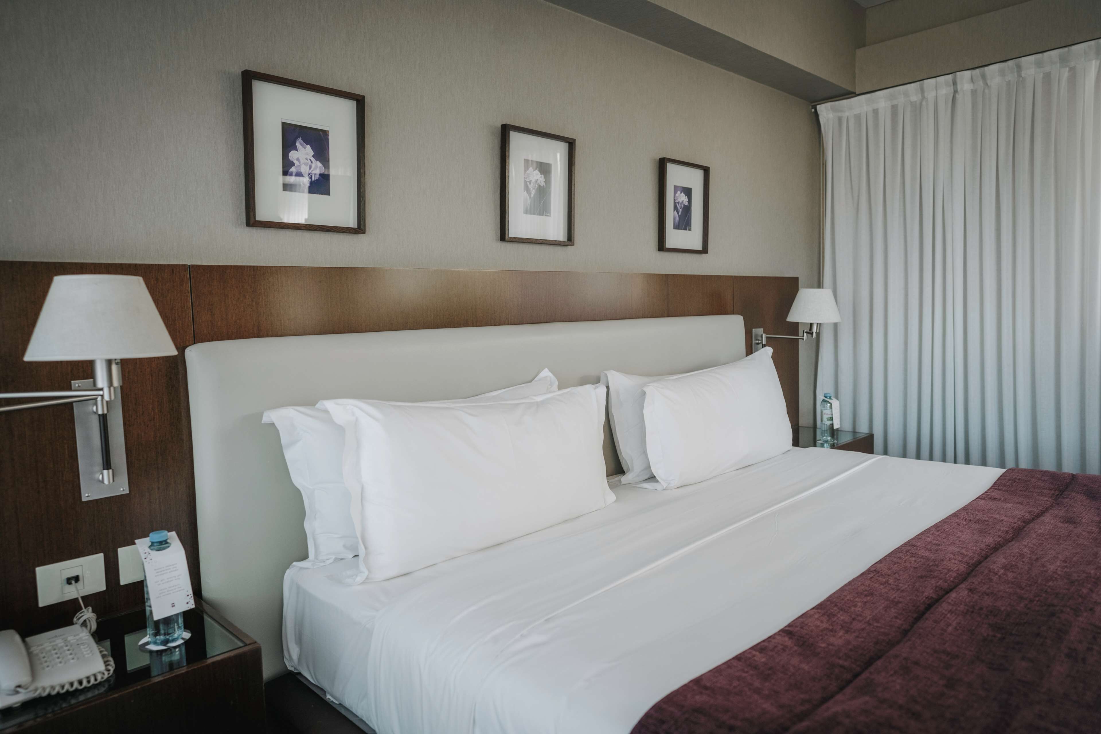 Une chambre d’hôtel avec un grand lit, deux lampes de chevet, un téléphone et trois tableaux encadrés accrochés au mur.