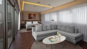 Een moderne hotelkamer met een grijze modulaire bank, een ronde salontafel en een groot raam met dunne gordijnen.