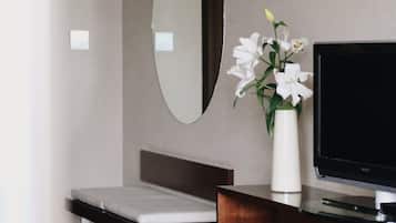 A modern hotel room with a wooden desk, a flat-screen TV, a large mirror, and a vase with flowers.