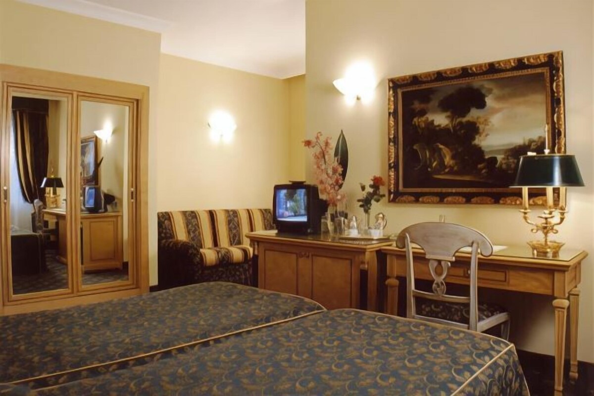 A hotel room with two beds, a desk with a television, a chair, a desk lamp, and a painting on the wall.