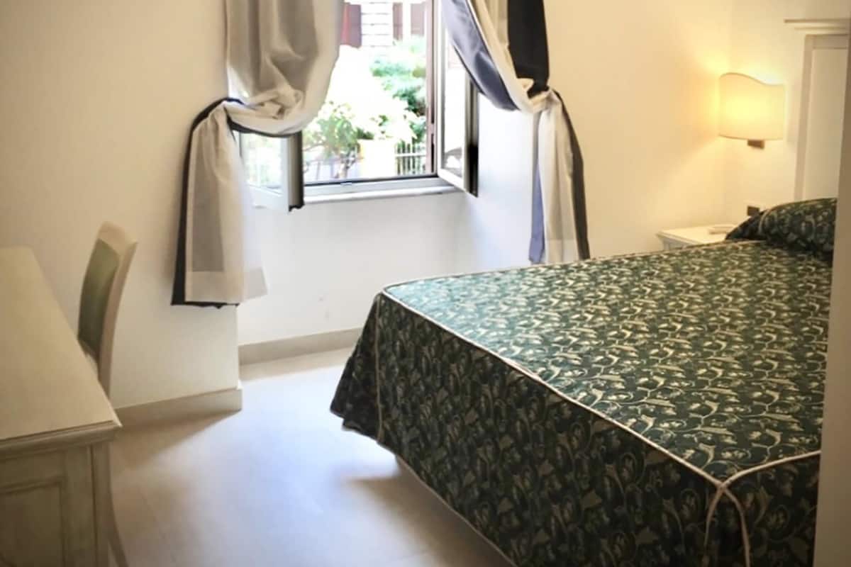 Une chambre à coucher avec un lit, une fenêtre avec des rideaux, une chaise et une table de chevet.