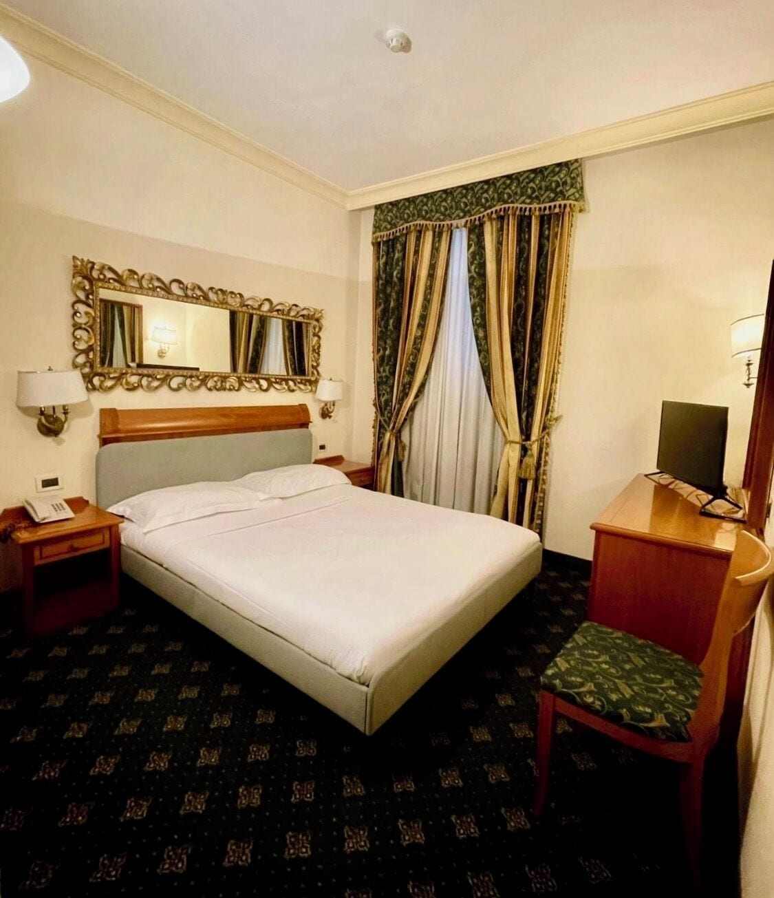 Habitación de hotel con cama, escritorio, silla, espejo, lámpara y televisor.